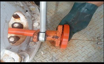 Fotografia de um operador com luva de proteção usando o torquímetro acoplado ao adaptador para apertar a tubulação na válvula angular de um tanque, sobre piso de chapa xadrez